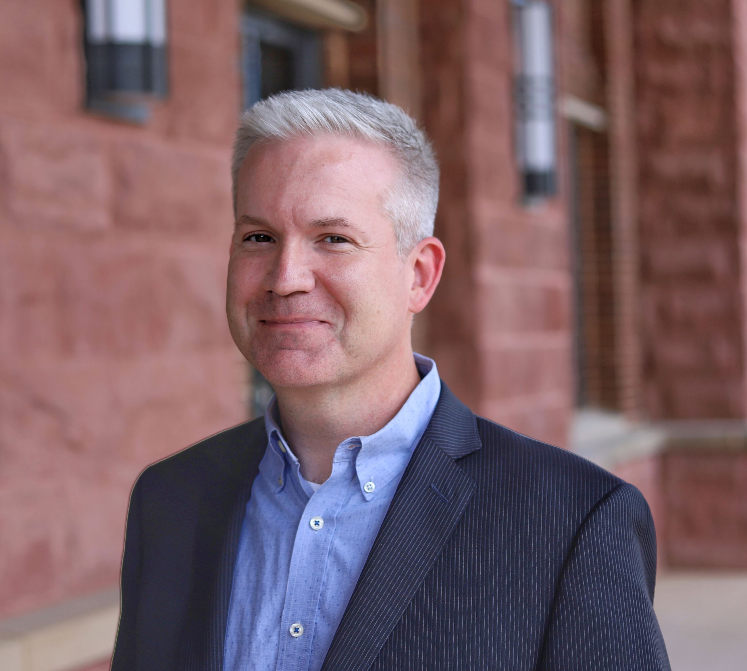 man with short gray hair smiling wearing a ligh blue shirt and dark sport coat in front of brick 
