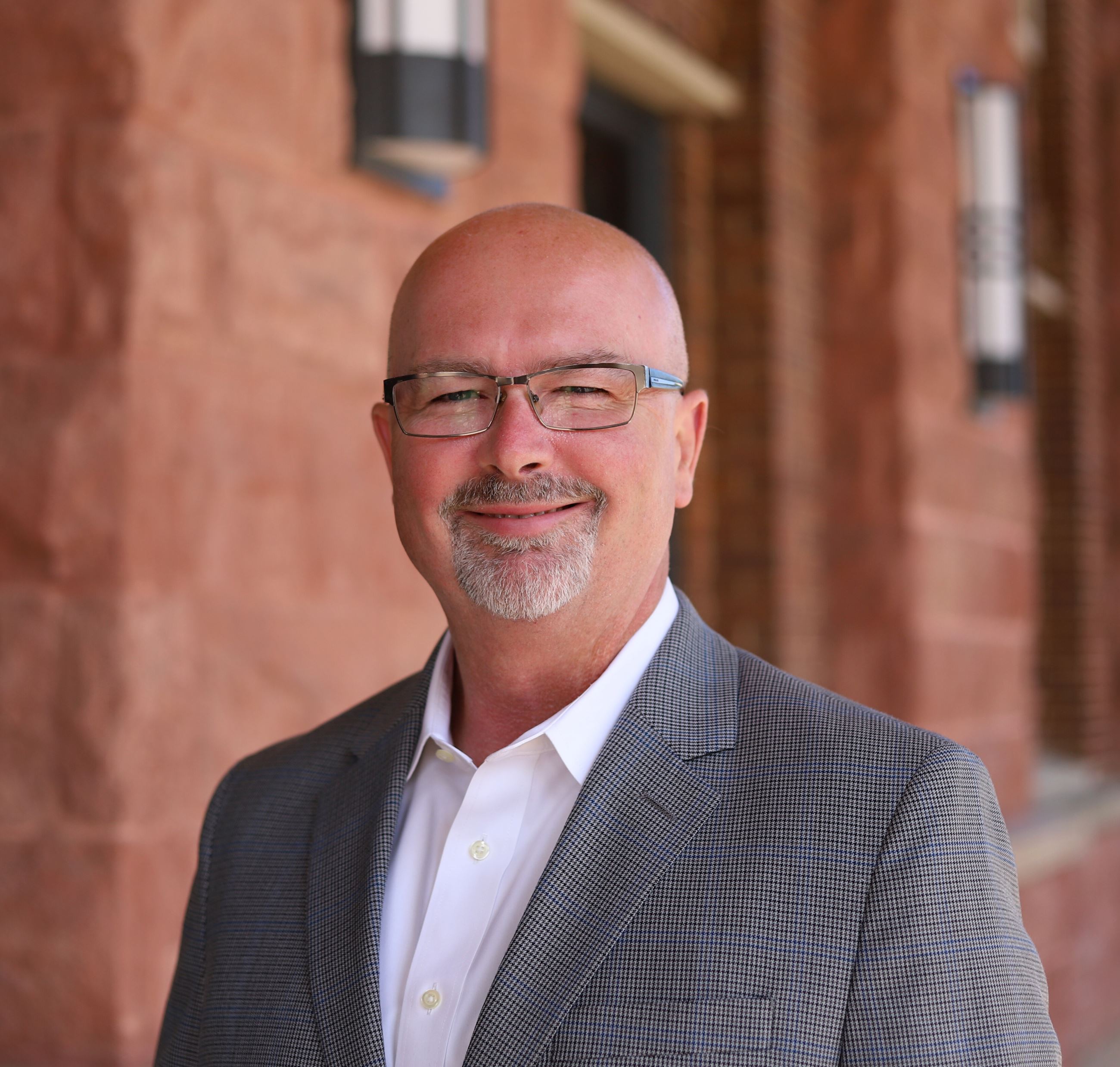 bald man wearing glasses, white shirt, sport coat smiling in front of brick building