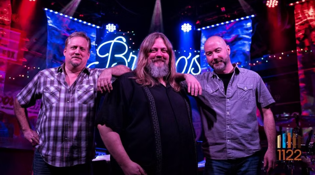 Three males standing in a dark room with accent lighting in the background.
